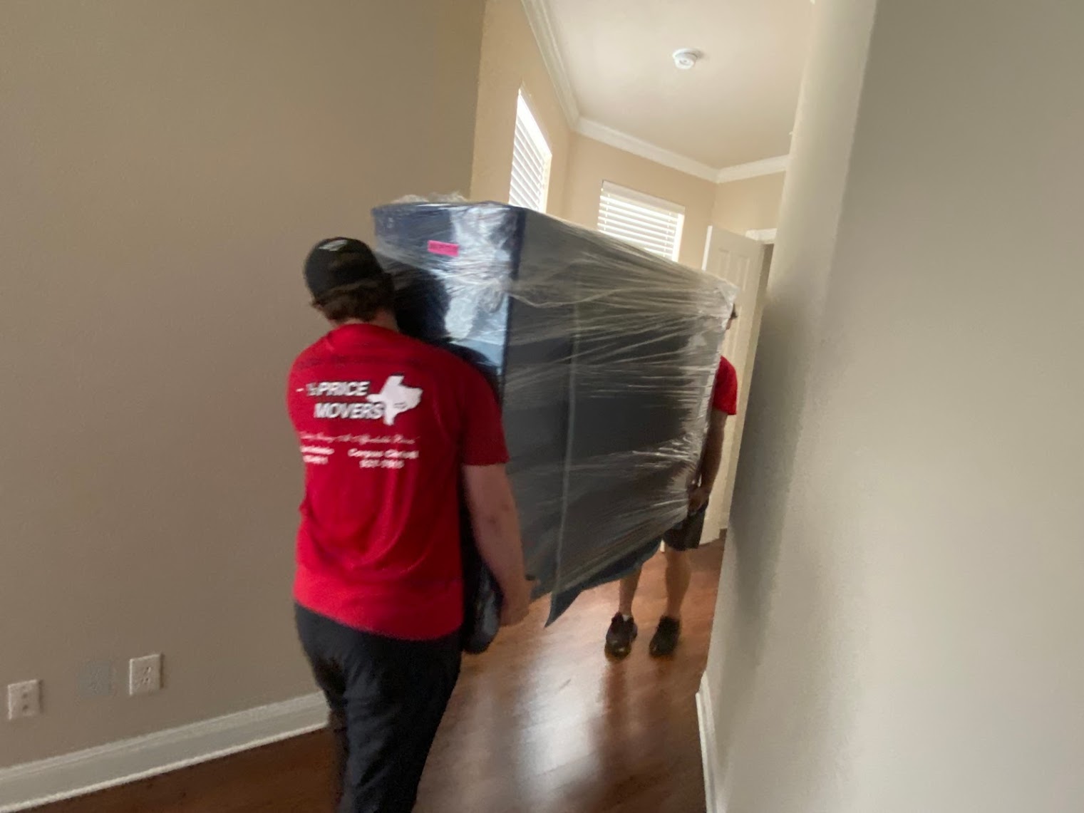 Two Half Price Movers crew members carrying wrapped furniture down an apartment hallway